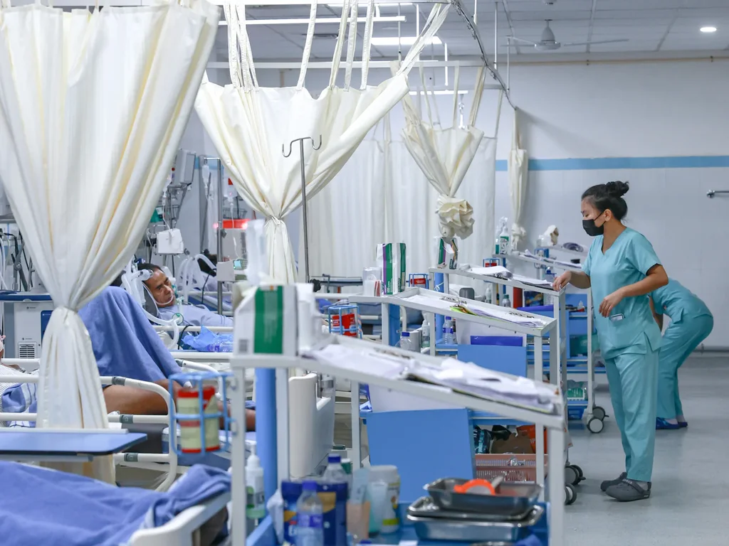 In-patient ward: patients resting in curtained beds while a nurse in scrubs checks the charts on a bedside trolley